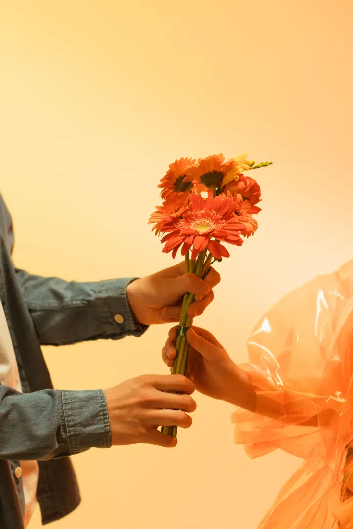 Una mano entrega gerberas naranjas a otra sobre un fondo cálido